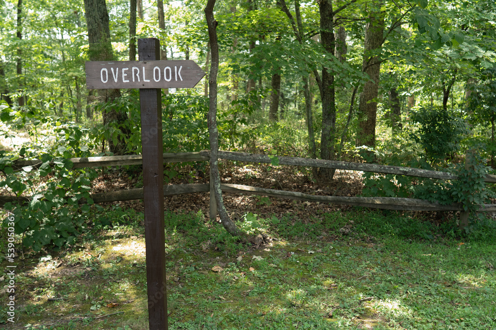 Wooden arrow sign that reads overlook and points to the right, set on a wooden post in the grass along a fence that runs with the hiking trail to the mountain view site Wooden arrow sign that reads overlook and points to the right, set on a wooden post in the grass along a fence that runs with the hiking trail to the mountain view site