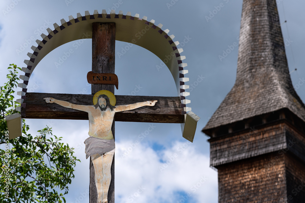 Old cemetery traditional cross depicting Jesus with a wood church in the background in Maramures region of Romania. INRI refers to “Iesus Nazarenus, Rex Iudaeorum,” meaning “Jesus of Nazareth, King of