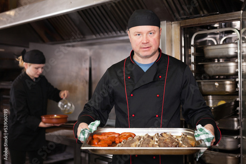 Skilled cook presenting appetizing baked pork ribs and sausages in restaurant kitchen, ready to serve it to guests
