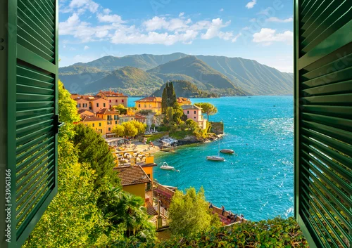 Naklejka na ścianę View through an open window with shutters looking down on the colorful picturesque village of Varenna, Italy, on the shores of Lake Como at summer.