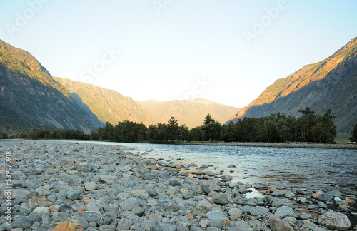 A shallow river with rocky banks in the shadow of the mountains from the setting sun in the autumn evening.