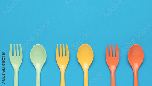 Orange, yellow and green plastic forks and spoons on a blue background. Minimal concept of plastic tableware.