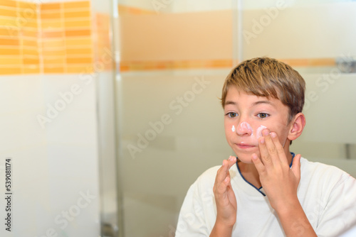 A preteen puts moisturizer on her face with her hands in front of the bathroom mirror.