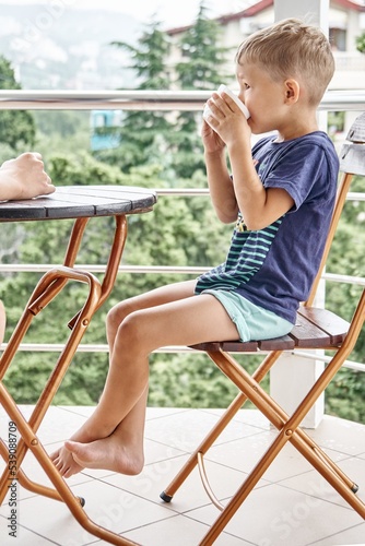 Preschooler boy drinks warm milk from mug sitting on chair in early morning. Excited child enjoys summer holidays drinking beverage on hotel balcony
