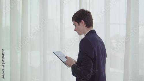 Young businessman walking and using tablet with white curtain in hotel. Caucasian man wear black suit