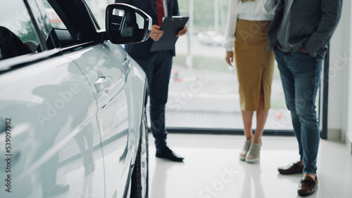 Low shot of prosperous people young couple buying new car in dealership and talking to helpful manager in suit. Shiny luxurious car is in foreground, large window is in background.