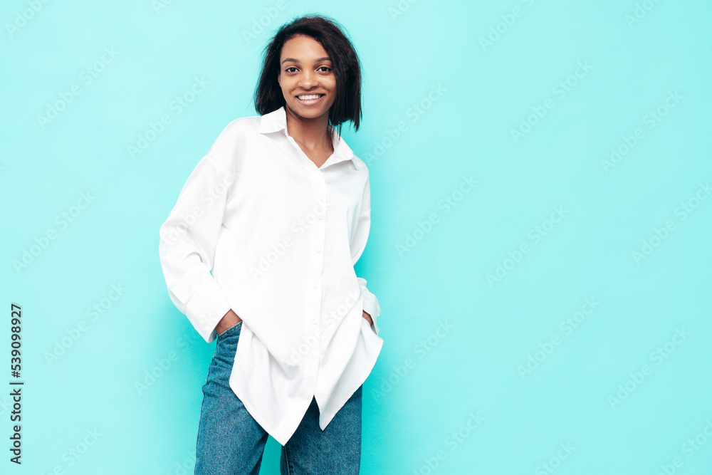 Portrait of young beautiful black woman. Smiling model dressed in summer jeans clothes. Sexy carefree female posing near blue wall in studio. Tanned and cheerful