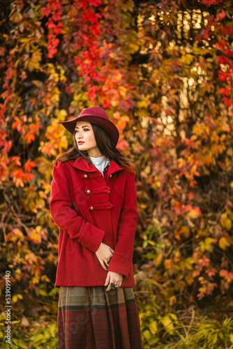 Beautiful young woman in an elegant vintage outfit and a red hat against the background of wild grapes  autumn  October