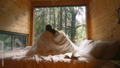 Happy couple sitting in bed covered by white blanket, hugging, kissing. Beautiful romantic middle aged husband and wife relaxing having fun together at forest cabin window, enjoy care love tenderness