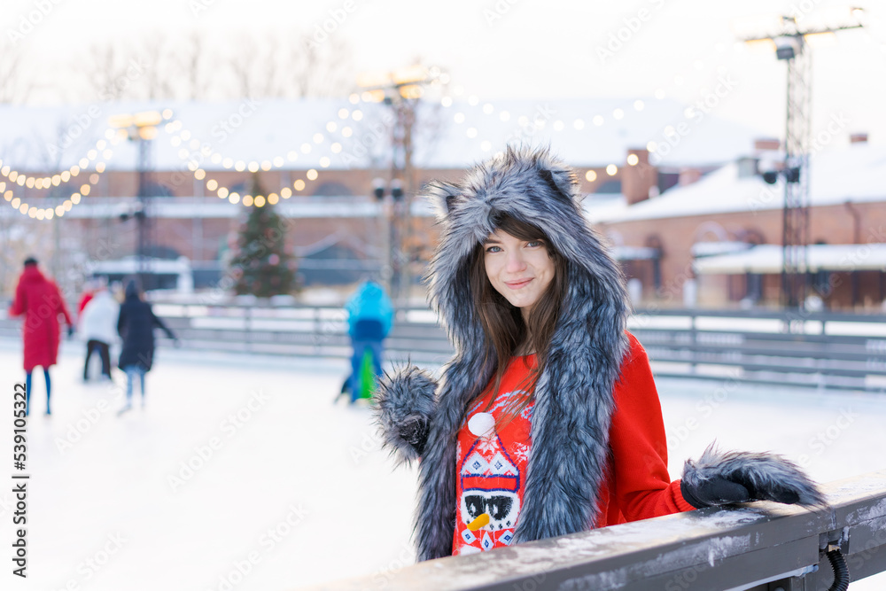 Portrait young woman in fur hat with ears and red sweater, with gloves on ice rinks, outdoors in winter. Happy woman is engaged in winter sport, entertainment on christmas holidays