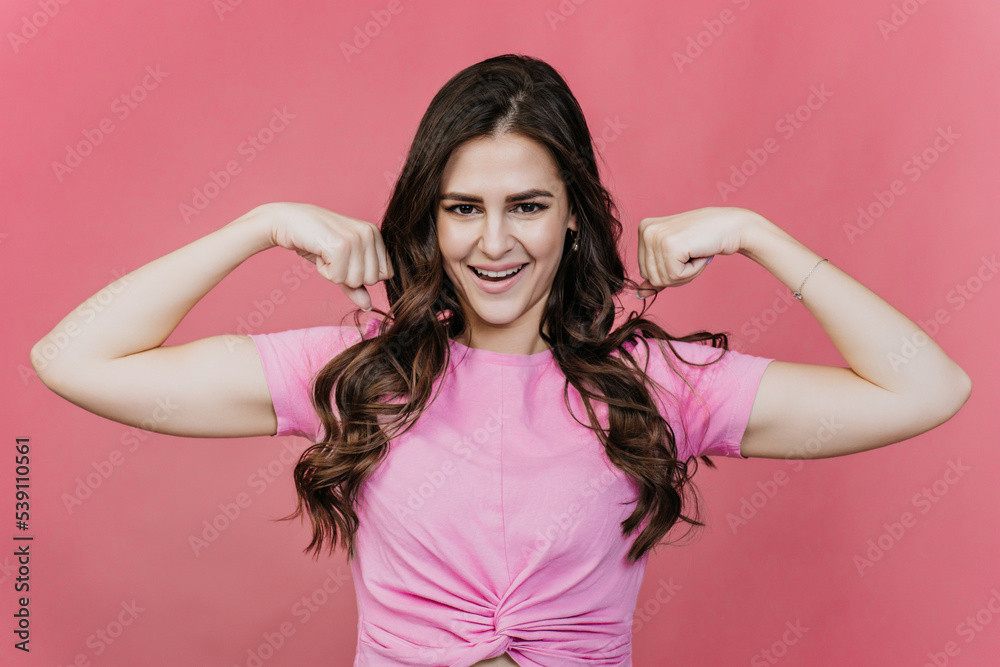 Fit hispanic young woman with loose wavy hair shows her biceps over pink studio backdrop. Attractive cheerful brunette girl in pink t-shirt at studio in perfect shape. Fitness, healthy lifestyle. Fit hispanic young woman with loose wavy hair shows her biceps over pink studio backdrop. Attractive cheerful brunette girl in pink t-shirt at studio in perfect shape. Fitness, healthy lifestyle.