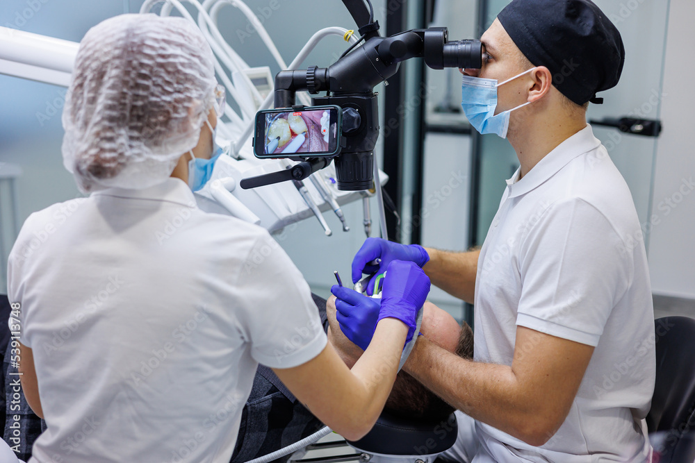 A dentist and an assistant treat a patient's teeth using a microscope. Professional teeth cleaning. Dental office. Dental microscope. A dentist and an assistant treat a patient's teeth using a microscope. Professional teeth cleaning. Dental office. Dental microscope.