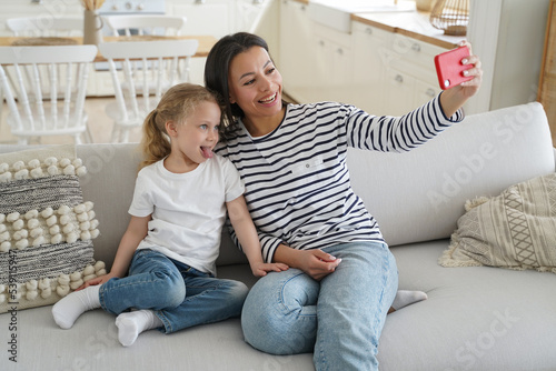 Mother, adopted little daughter, showing tongue, take family selfie together, using phone on couch