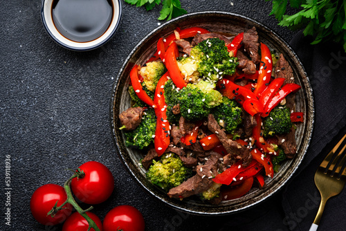 Stir fry vegetables with beef, paprika and broccoli with sesame seeds in bowl on black table  background, top view