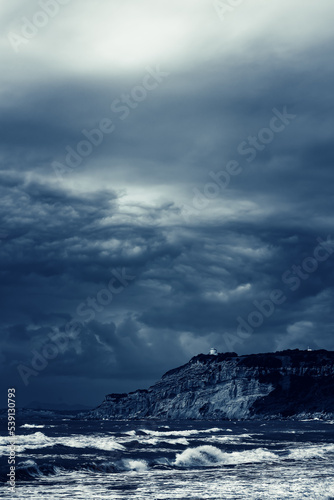 Turbulent ocean with a small house on a cliff under a dark cloudy sky.