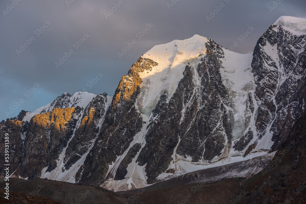 Gloomy landscape with giant snowy mountain top and sunlit gold rocks in dramatic gray sky. Huge snow mountains with hanging glacier and cornice under rainy clouds. Snow-covered mountains in overcast.