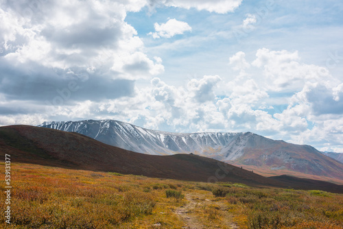 Motley autumn landscape with hills on high plateau and sunlit snowy mountain range under dramatic cloudy sky. Vivid autumn colors in mountains. Sunlight and shadows of clouds in changeable weather.