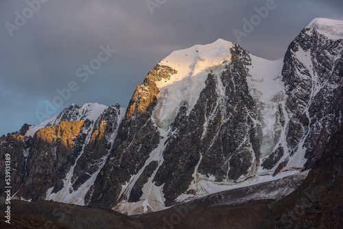 Gloomy landscape with giant snowy mountain top and sunlit gold rocks in dramatic gray sky. Huge snow mountains with hanging glacier and cornice under rainy clouds. Snow-covered mountains in overcast. © Daniil