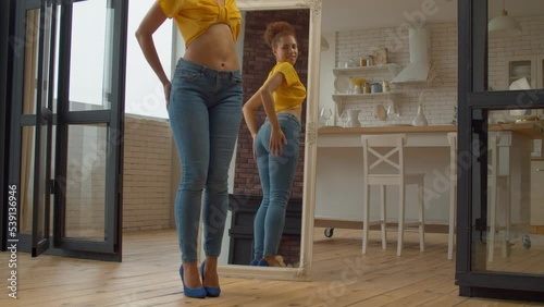 Joyful attractive African American woman with perfect body in stylish clothes and high heels looking at reflection in mirror, admiring appearance and shapes after successful weight loss at home. photo