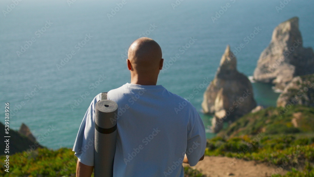 Fototapeta premium Yoga man walking edge cliff holding mat. Focused sportsman looking ocean coast