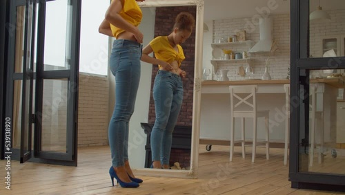 Reflection in mirror of upset charming African American female in stylish clothes disappointed by her appearance and shapes, feeling fat after gaining weight in domestic room,