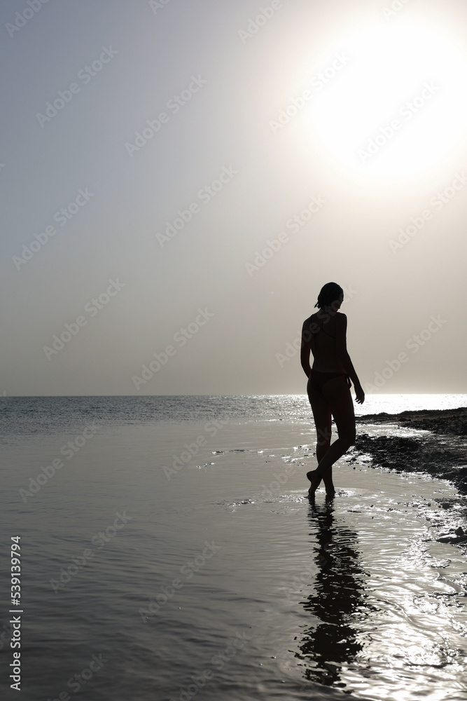 woman silhouette in bikini walking in the water at sunset on the sea shore