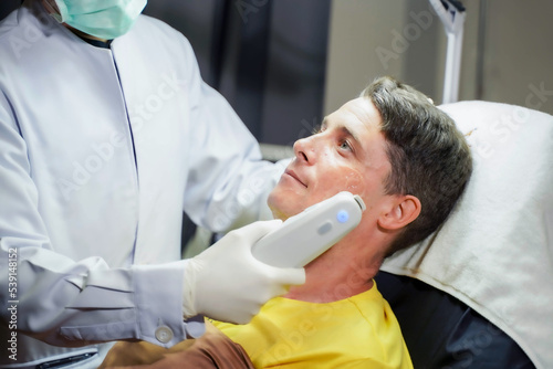 Closeup handsome man having color light therapy to stimulate facial skin by professional cosmetologist wellbeing.