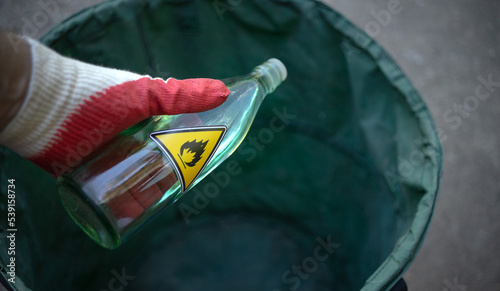 A gloved hand holds a bottle of flammable liquid. Disposal of flammable substances. The release of gorenje products to the trash. Yellow triangular sign of dangerous liquid in the bottle.