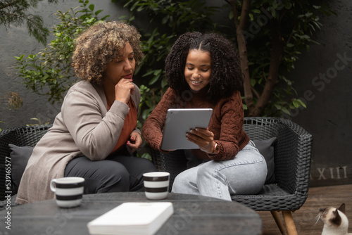An African-American woman and her daughter use a digital tablet for online shopping. They sit on the patio and drink a coffee. The daughter is petting her siamese cat.