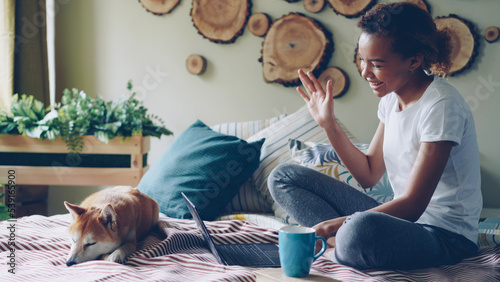 Happy girl is skyping talking online with friends gesturing and looking at laptop screen sitting on bed at home with cute puppy lying beside her. Communication and technology concept. photo