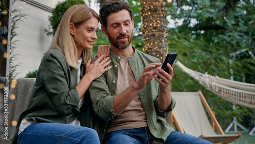 Married couple husband and wife Caucasian bearded man and blond woman 40s spouses outdoor in backyard at campsite looking at phone choose buy online booking with smartphone using mobile e-shopping app © Yuliia