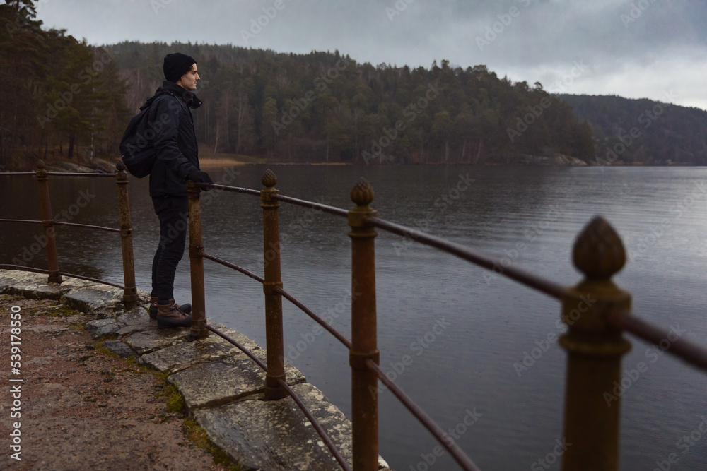 A caucasian man with a backpack standing by a old fence looking out over a lake at a rainy day.