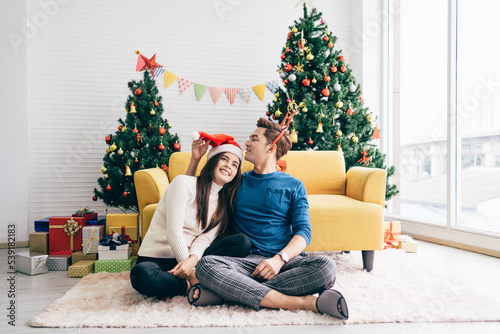 Young beautiful happy Asian woman wearing a Santa Claus hat sitting together with her boyfriend at home with a Christmas tree in the background. Image with copy space.