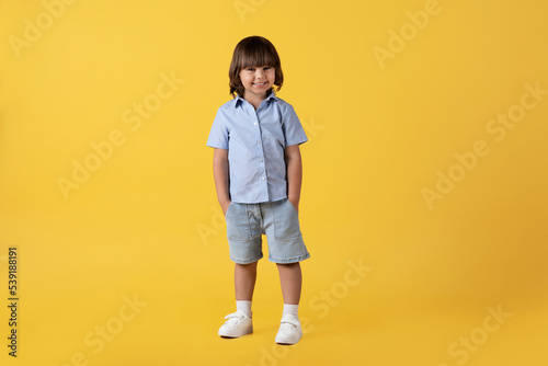 Full length portrait of adorable carefree little boy smiling to camera, holding hands in pockets, orange background © Prostock-studio
