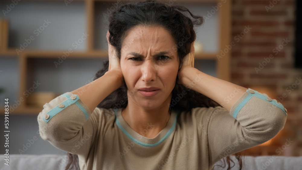 Portrait Hispanic Caucasian woman irritated distraught girl sitting at home covers ears with hands ignores refuses to listen hear protects from noise shouting loud sounds holds head suffers headache Portrait Hispanic Caucasian woman irritated distraught girl sitting at home covers ears with hands ignores refuses to listen hear protects from noise shouting loud sounds holds head suffers headache