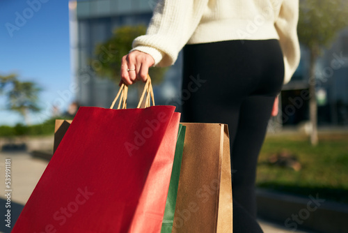 Close up of woman walking with shopping bags in hands in city from mall. Low angle view of girl going with purchases from black friday sale. Female with full paper bags. Back view.