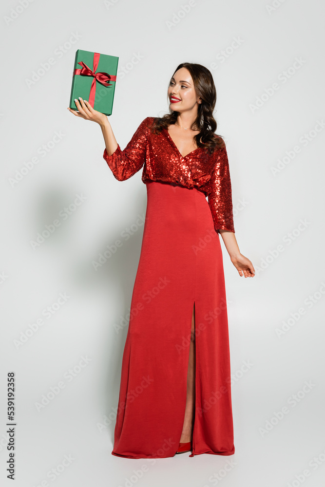 Fototapeta premium Full length of stylish woman in red dress looking at gift box on grey background
