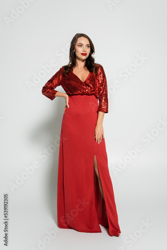 Full length of pretty brunette woman in red dress holding hand on hip on grey background