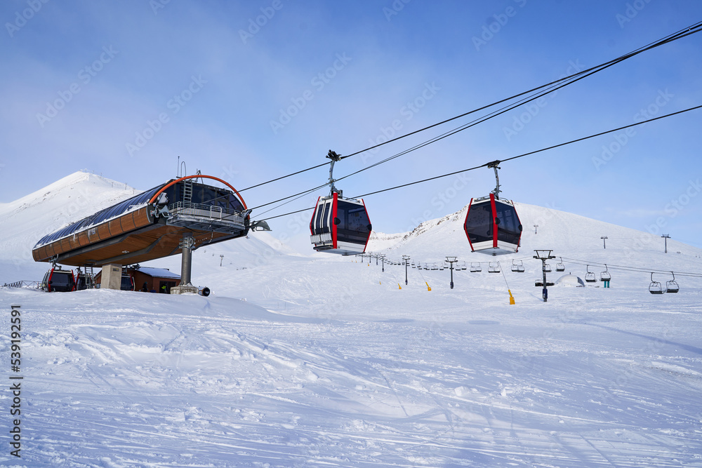 Naklejka premium Cable car gondola at ski resort with snowy mountains on background. Modern ski lift with funitels and supporting towers high in the mountains on winter day. Ski lift station with no people.