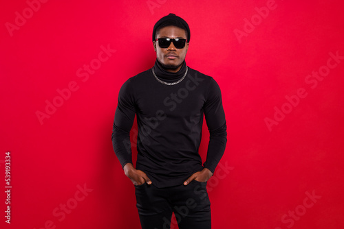 Portrait of attractive cool serious guy holding hands in pockets wearing black look isolated over bright red color background