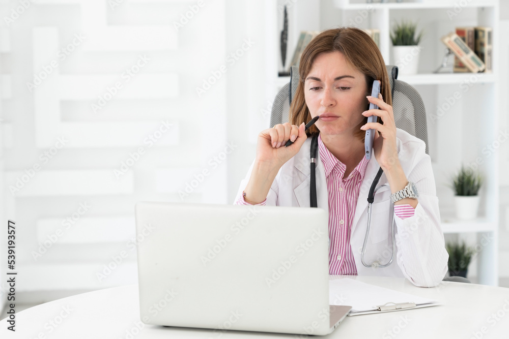 Portrait photo of young and beautiful serious female doctor in white medical gown sitting and working on laptop in the office of the modern clinic. Online consultation and distant cure concept