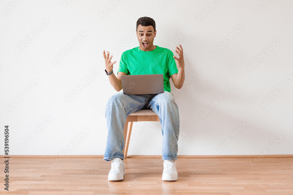 Online offer. Emotional arab man using laptop and shouting, celebrating news while sitting on chair and spreading hands