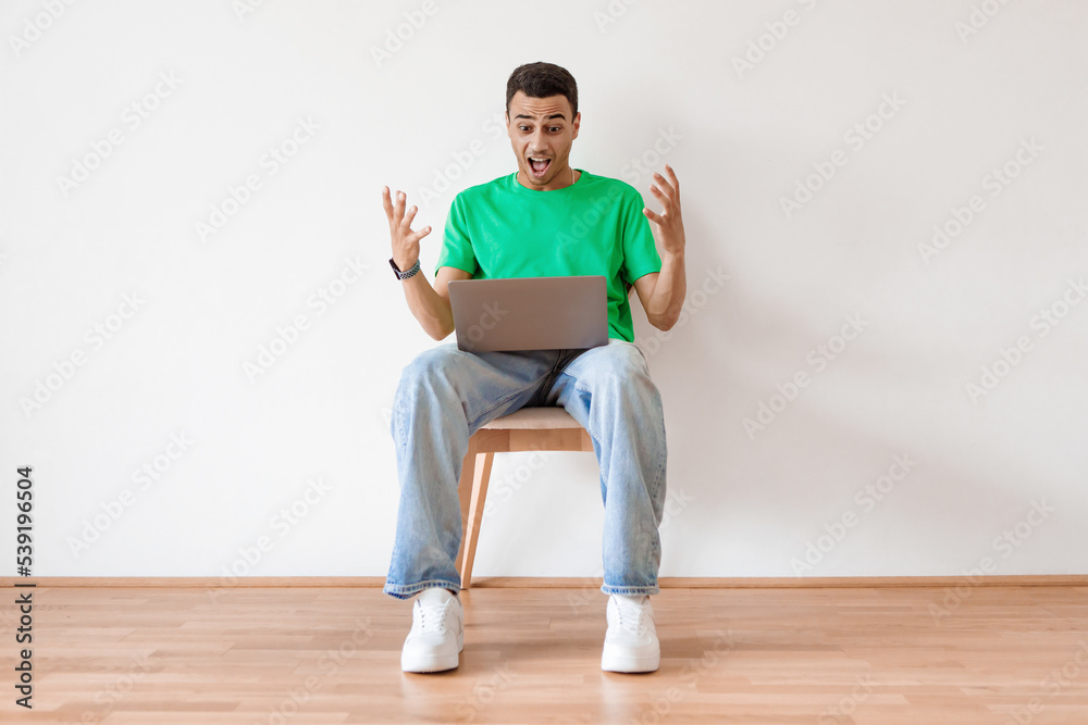 Online offer. Emotional arab man using laptop and shouting, celebrating news while sitting on chair and spreading hands Online offer. Emotional arab man using laptop and shouting, celebrating news while sitting on chair and spreading hands
