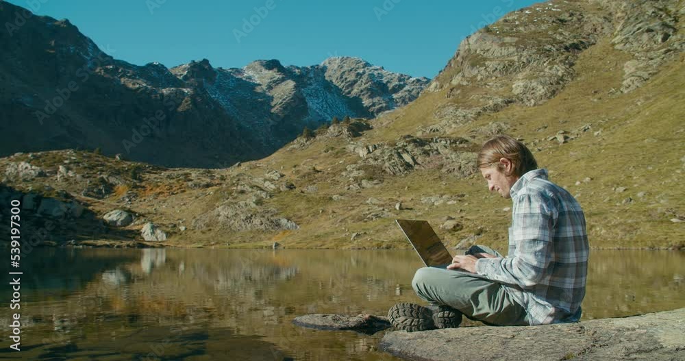 Man sit on mountain lake shore using laptop computer on vacation trip adventure Man sit on mountain lake shore using laptop computer on vacation trip adventure