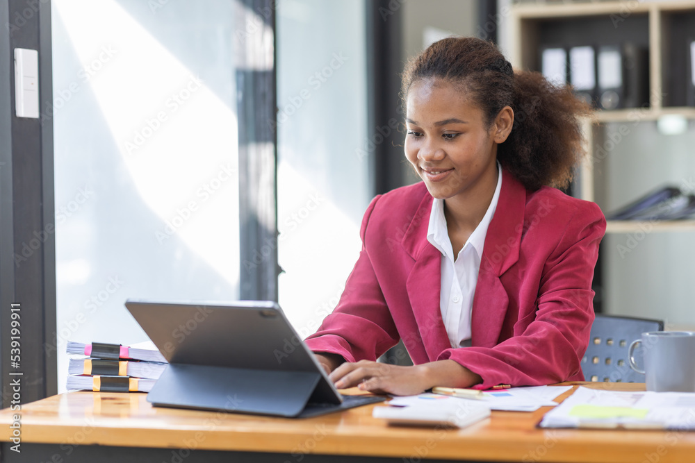Smiling young African american female Finance Entrepreneur sitting in workplace office reading paperwork and working on a laptop, investment financial document report finance analysis concept. Smiling young African american female Finance Entrepreneur sitting in workplace office reading paperwork and working on a laptop, investment financial document report finance analysis concept.