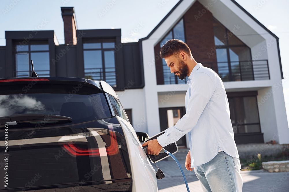 Process of charging the vehicle. Young stylish man is with electric car at daytime Process of charging the vehicle. Young stylish man is with electric car at daytime