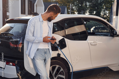 Beautiful young stylish man is with electric car at daytime charging the vehicle and holding smartphone © standret