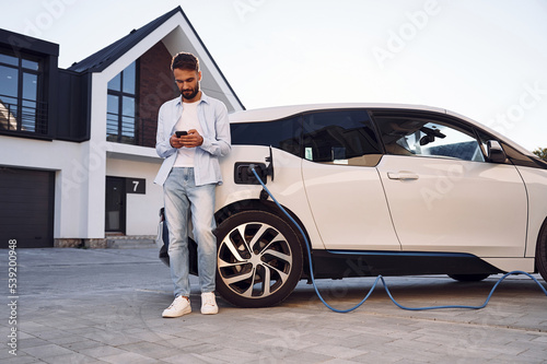 Beautiful young stylish man is with electric car at daytime charging the vehicle and holding smartphone © standret