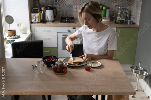 Mature woman sitting at table in kitchen makes breakfast. Daylight breaks through window illuminating small kitchen. Middle aged female spreads butter on toasts and makes tea in cozy kitchen