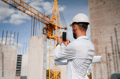 Taking a picture of place by smartphone. Man in uniform is working on the construction site